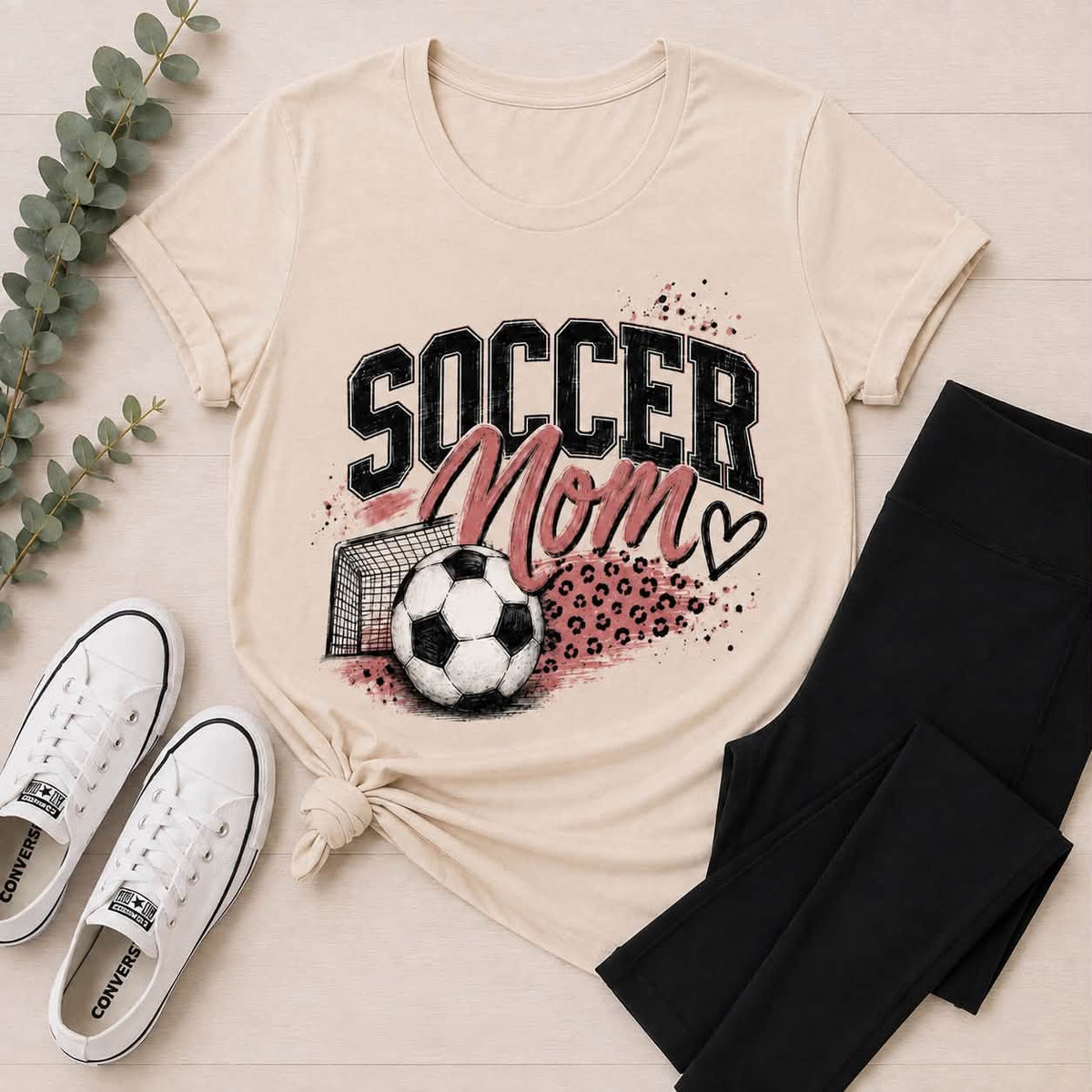 Cream crewneck tee featuring a black and pink Soccer Mom graphic with a soccer ball, goal, leopard print accents, and heart.