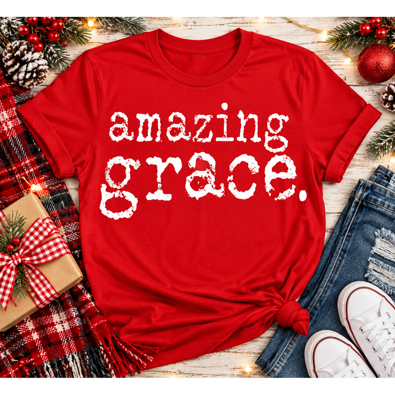 A red short-sleeve graphic T-shirt with the phrase 'amazing grace.' in distressed white lettering, styled in a holiday flat lay with jeans, white sneakers, a plaid blanket, pinecones, and Christmas decorations.