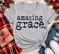 A heather gray short-sleeve t-shirt with 'amazing grace.' printed in black distressed text across the front, styled in a flat lay with jeans, white sneakers, a red plaid scarf, pinecones, and holiday decorations.