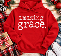 A red pullover hoodie with the words 'amazing grace.' printed in white distressed lettering across the chest, styled in a holiday flat-lay with a plaid scarf, gift box, pinecones, ornaments, jeans, and white sneakers on a wooden surface.
