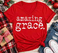 A red V-neck t-shirt with the words 'amazing grace.' printed in a distressed white font, styled in a flat lay with jeans, white sneakers, a plaid scarf, a wrapped gift, pinecones, and holiday berries on a light wood background.