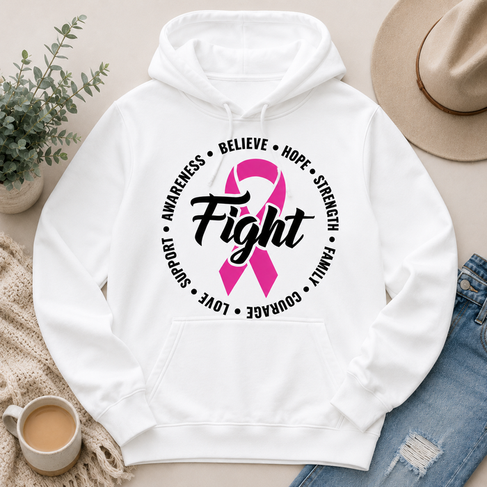 A white pullover hoodie laid flat with a pink awareness ribbon and the word 'Fight' printed on the chest, surrounded by black text reading 'Believe', 'Hope', 'Strength', 'Family', 'Love', 'Courage', 'Support', and 'Awareness'. The hoodie is styled in a flat lay with a hat, jeans, a plant, a cup of coffee, and a knit blanket on a light beige surface.