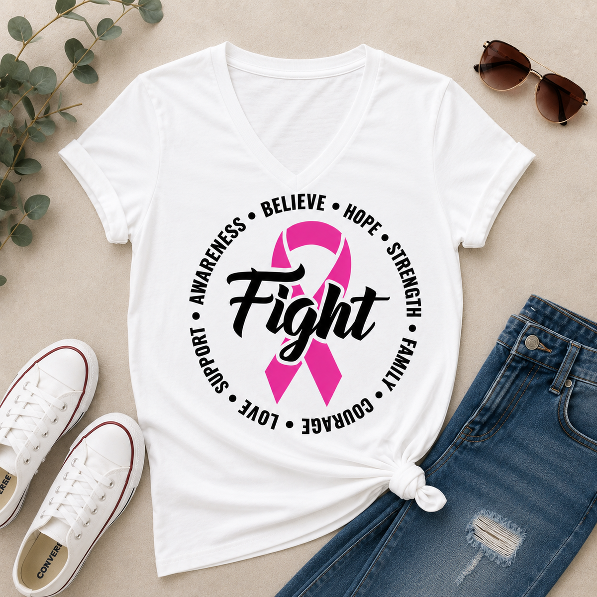 White short-sleeve V-neck T-shirt laid flat with a pink ribbon and the word 'Fight' printed on the front, surrounded by black text reading 'Awareness • Believe • Hope • Strength • Family • Love • Courage'. Styled flat-lay with blue jeans, white sneakers, sunglasses, and eucalyptus leaves on a beige surface.