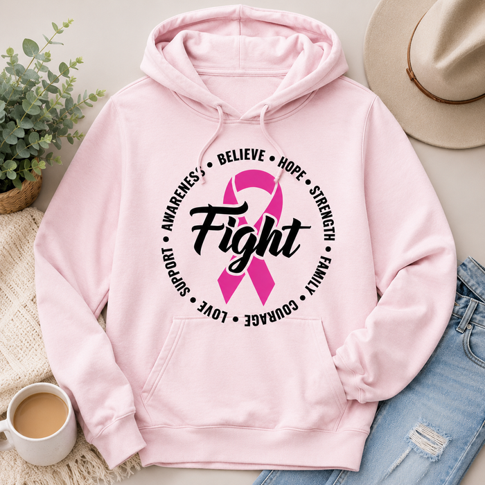 A light pink pullover hoodie with a pink awareness ribbon and the word 'Fight' printed on the chest, styled in a flat lay with a hat, jeans, a plant, a mug, and a blanket.