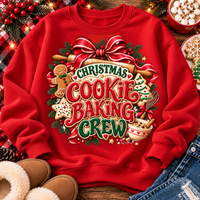 A red crewneck sweatshirt laid flat on a rustic wooden surface, featuring a large festive graphic that reads 'Christmas Cookie Baking Crew' with holiday illustrations including a gingerbread man, cookie, tree, ornaments, and baking tools. The sweatshirt is styled with holiday greenery, lights, boots, denim, and dessert props around it.