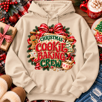 A beige pullover hoodie with a large Christmas cookie baking crew graphic, styled in a cozy holiday flat-lay with a gift box, slippers, denim, and a mug of hot chocolate.