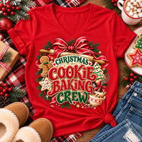 A red short-sleeve V-neck T-shirt laid flat on a rustic holiday surface, featuring a large Christmas cookie baking graphic with the words “CHRISTMAS COOKIE BAKING CREW,” cookies, gingerbread, ornaments, pinecones, and a red bow. Surrounding props include jeans, slippers, a mug of hot cocoa with marshmallows, wrapped gift boxes, and cookies.
