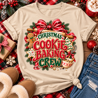 A beige graphic t-shirt with a festive Christmas cookie baking crew design, styled in a flat lay with holiday decorations, slippers, jeans, and a mug of popcorn.