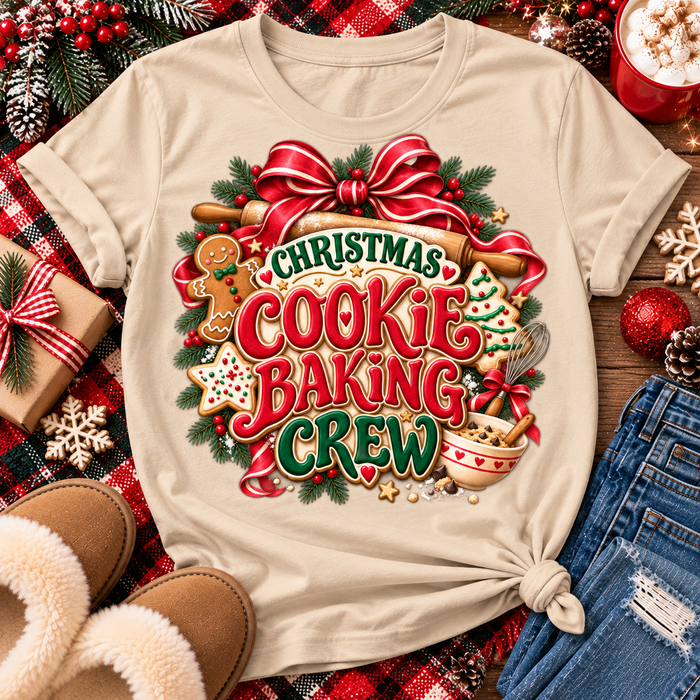 A beige graphic t-shirt with a festive Christmas cookie baking crew design, styled in a flat lay with holiday decorations, slippers, jeans, and a mug of popcorn.