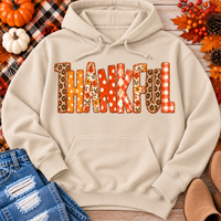 Beige Thankful fall graphic hoodie with orange floral, plaid and polka dot patchwork lettering, styled with jeans and autumn decorations.