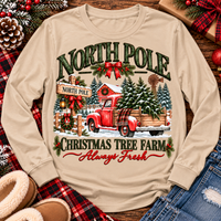 North Pole Christmas Tree Farm vintage holiday graphic shirt featuring a red pickup truck carrying Christmas trees, snowy evergreen trees, rustic farmhouse scenery, festive red bows, and distressed green lettering reading North Pole Christmas Tree Farm Always Fresh. Available in beige and black in short sleeve crewneck, V-neck, long sleeve crewneck, and hoodie styles.