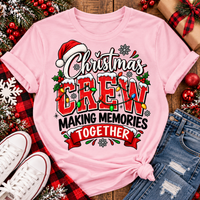 Christmas Crew Making Memories Together graphic on a pink crewneck T-shirt, featuring red lettering, colorful Christmas lights, a Santa hat, holly, and snowflakes.