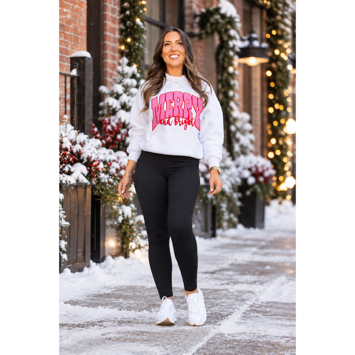 A woman stands outdoors in a snowy walkway wearing a white crewneck sweatshirt with a pink 'Merry & Bright' graphic, black leggings, and white sneakers.