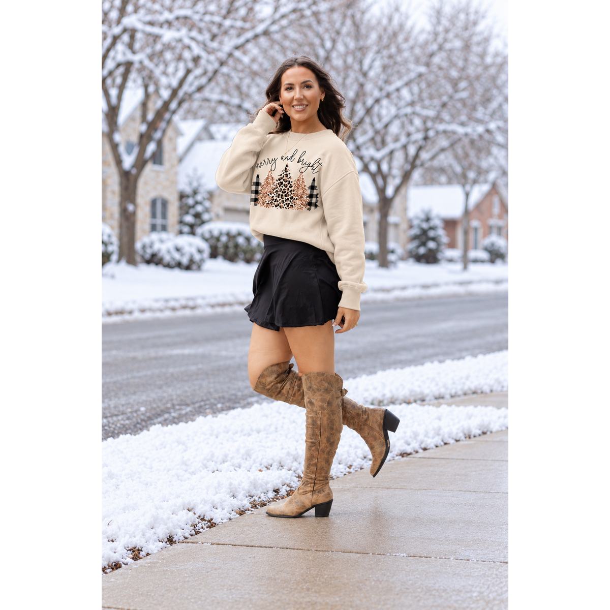 A woman stands on a snowy sidewalk wearing a cream crewneck sweatshirt with a Christmas tree graphic, a black mini skirt, and brown knee-high boots.
