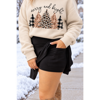 A person wearing a cream sweatshirt with a 'merry and bright' holiday tree graphic in leopard print, glitter, and plaid, paired with black shorts in a snowy outdoor setting.