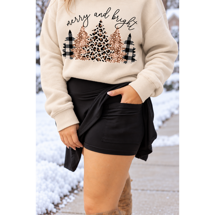 A person wearing a cream sweatshirt with a 'merry and bright' holiday tree graphic in leopard print, glitter, and plaid, paired with black shorts in a snowy outdoor setting.