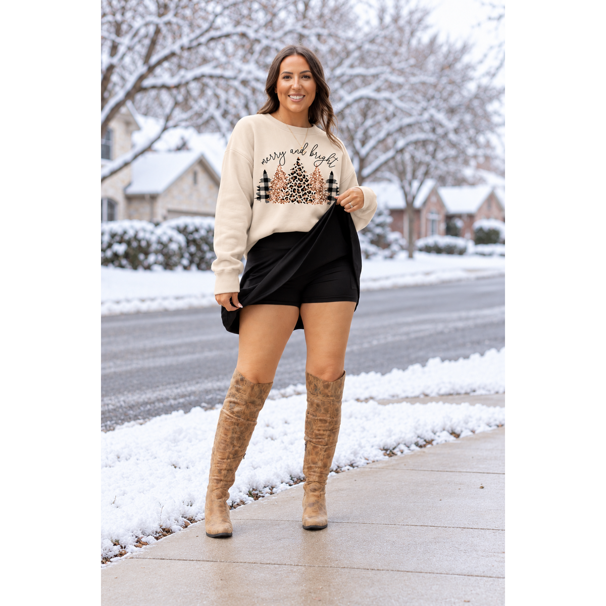 A woman stands on a snowy sidewalk wearing a cream crewneck sweatshirt with a 'Merry and Bright' Christmas tree graphic, a black mini skirt, and brown knee-high boots.