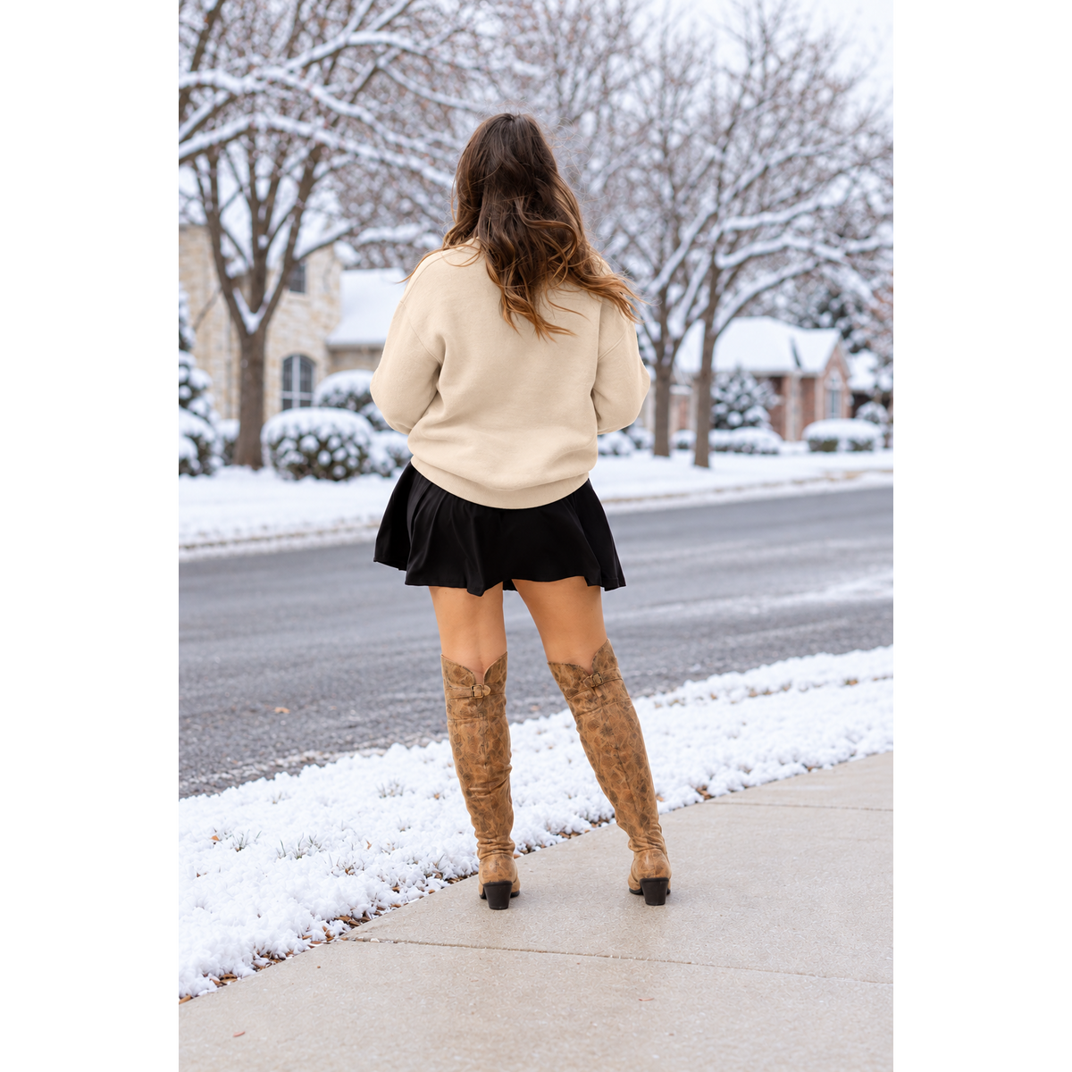 A woman stands on a snowy sidewalk, viewed from behind, wearing a cream knit sweater, a black pleated mini skirt, and brown faux-suede knee-high boots with western-style cutouts. The background shows a residential street with snow-covered trees and houses.