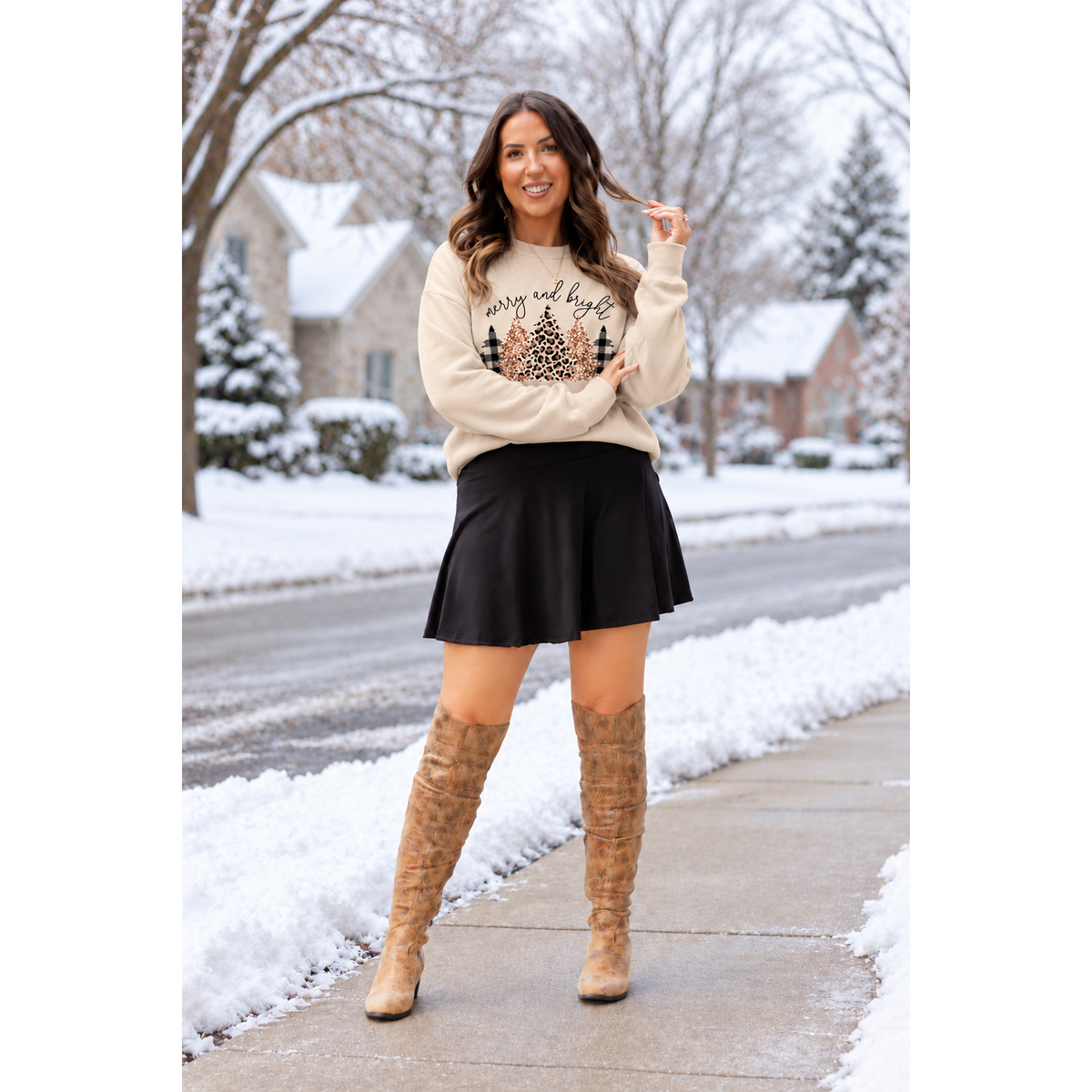 A woman stands on a snowy sidewalk wearing a cream crewneck sweatshirt with a holiday tree graphic and the words 'Merry and Bright', a black pleated mini skirt, and tan knee-high boots.