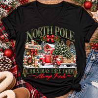 North Pole Christmas Tree Farm vintage holiday graphic shirt featuring a red pickup truck carrying Christmas trees, snowy evergreen trees, rustic farmhouse scenery, festive red bows, and distressed green lettering reading North Pole Christmas Tree Farm Always Fresh. Available in beige and black in short sleeve crewneck, V-neck, long sleeve crewneck, and hoodie styles.