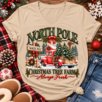 North Pole Christmas Tree Farm vintage holiday graphic shirt featuring a red pickup truck carrying Christmas trees, snowy evergreen trees, rustic farmhouse scenery, festive red bows, and distressed green lettering reading North Pole Christmas Tree Farm Always Fresh. Available in beige and black in short sleeve crewneck, V-neck, long sleeve crewneck, and hoodie styles.